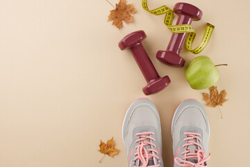 Fall fitness transformation idea. Top view composition of sneakers, tape measure, dumbbells, fresh apple, fallen leaves on pastel beige background with advertising zone
