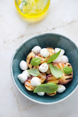 Grilled peaches with baby mozzarella and fresh basil in a green bowl, vertical shot on a light-beige marble background, high angle view