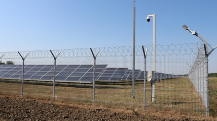 power station with solar panels in a field behind a barbed wire fence, lighting and video surveillance equipment on a bright sunny day, batteries accumulating solar energy in a countryside landscape