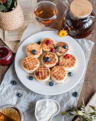 cheesecakes with a heap of raspberries and blueberries in a white plate, a golden fork and knife, tea in a glass cup and a kettle next to it, raspberry jam in a jar
