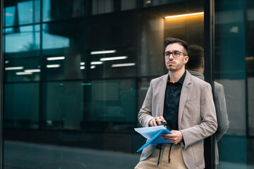 Overworked businessman standing in thoughts outside office building . Pensive man standing outdoors with paper documents and coffee to go.