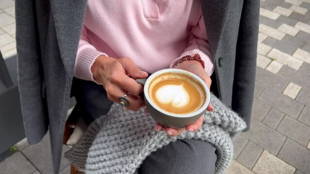 Woman sitting in outdoor cafe with cup of cappuccino coffee with heart in her hands comfortably wrapped in gray plaid scarf, Because it became chilly.