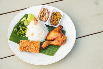 Cooked chicken meat with variety of side dish. Grilled chicken thigh with tumpeng white rice, fried tempeh, salty anchovy fish, lalapan vegetables, and sambal chili sauce. Hotel and restauran concept.