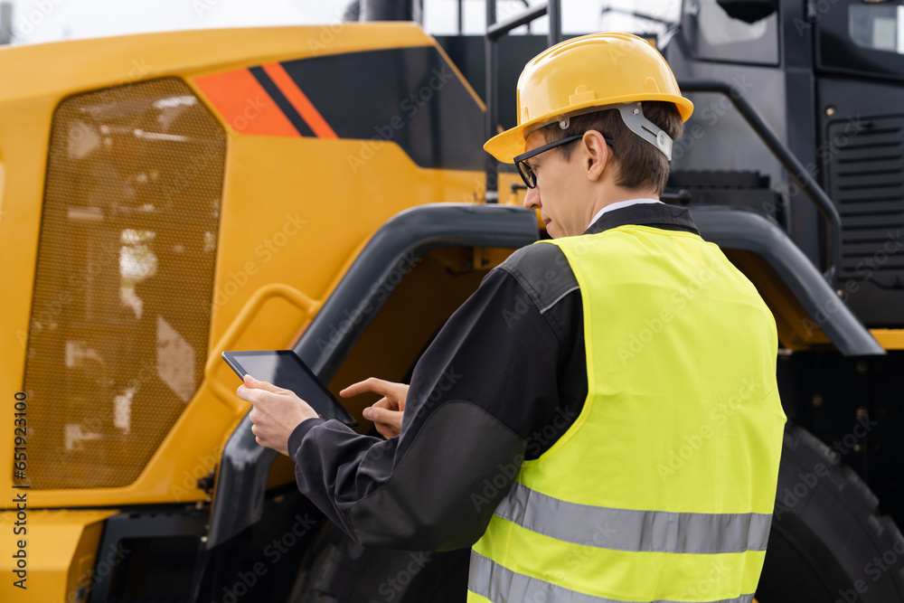 Poster Worker in a helmet with a digital tablet on the background of construction machine