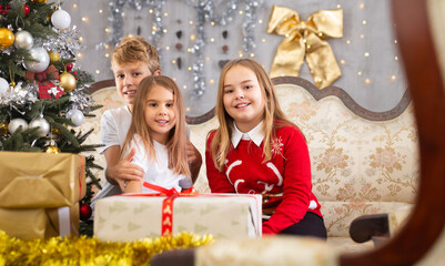 Fototapeta na wymiar Happy brother with two sisters are sitting on sofa together with gift boxes next to the new year tree