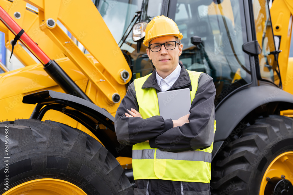 Poster Worker in a helmet with a digital tablet on the background of construction machine