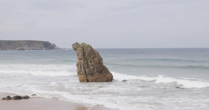 A scenic footage of a huge rock on a sandy beach with crashing waves under a majestic blue sky