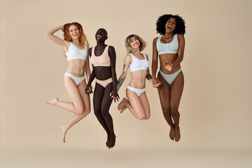 Four happy pretty diverse young women wearing underwear jumping on beige background. Positive multicultural ladies group, multiethnic girls friends models having fun, diversity and natural body beauty