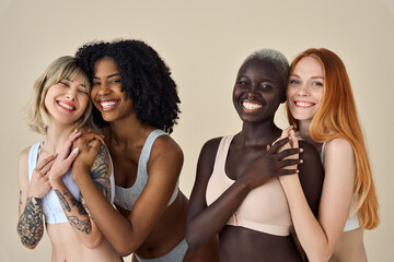 4 happy diverse women, four smiling multicultural young girls friends beauty International models faces group in underwear tops hugging looking at camera standing on beige background, portrait.