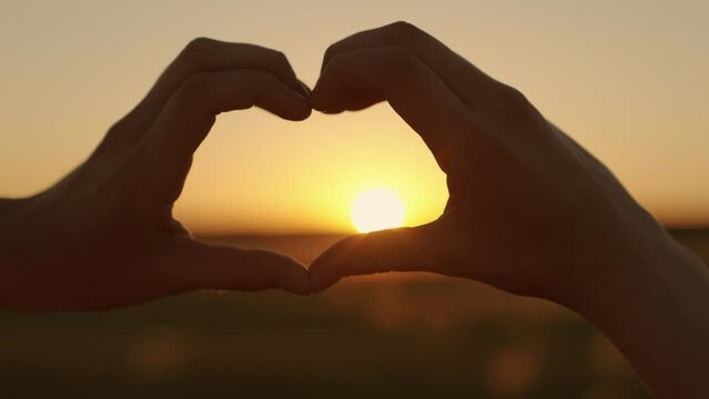 Heart shape made with fingers on background of sun. Silhouette of happy girl in park on sunset shows heart symbol with her fingers. Conception of health, love, freedom. Travel, nature. Childhood dream