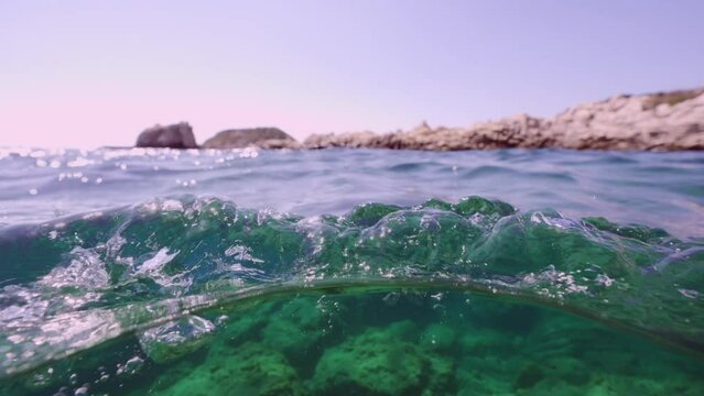 Split level, Camera moves forward at water level in rocky blue bay of Mediterranean Sea, Slow motion. Plastic pollution of Ocean