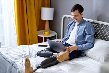 positive male with disability and special needs sit on bed and work on laptop. cheerful and excited. Handicapped Caucasian male using laptop for work or studies. New normal, social distancing