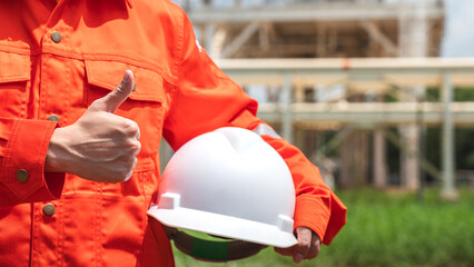 Action of the engineer in orange coverall uniform is holding a white safety helmet with background of chemical or petrochemical refinery plant. Ready to work in challenge job action concept.