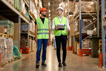 two engineers in safety helmets and green uniforms walking among shelves with goods in warehouse...