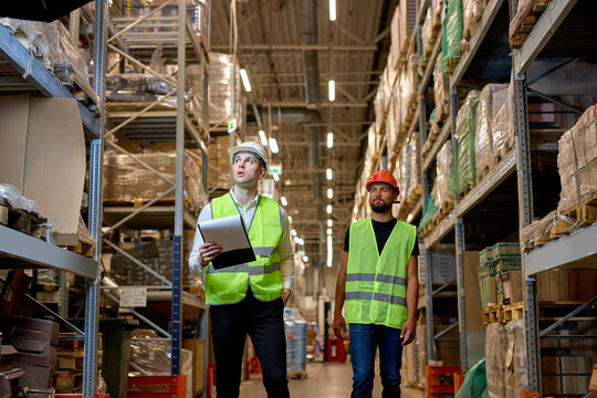 Retail Warehouse full of Shelves with Goods, Two Caucasian Male Workers Supervisors in Working Uniform and Helmet Discuss Product Delivery. Workers Walking and Having Talk In Warehouse