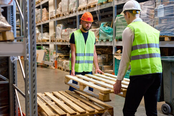 caucasian warehouse men hold cargo pallet together with co-worker in workplace area. hardworking strong guys in green vests working uniforms engaged in work in cargo distribution storage