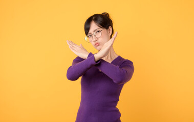 Explore concept of denial and rejection through X gesture made serious young woman wearing purple shirt and eyeglasses in studio portrait against a yellow background. An impactful symbol for caution.