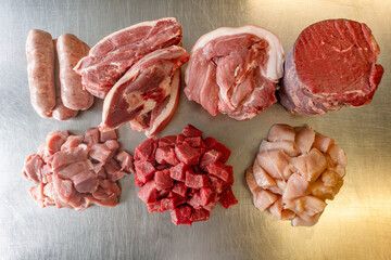 Selection of raw meat on a metal table: sausage, diced pork, lamb shoulder cuts, chopped beef, diced chicken breast, and rolled brisket.