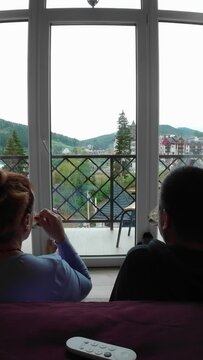 Man and Woman are sitting in front of the balcony window.in the hotel room He takes the remote control and swich day to evening.