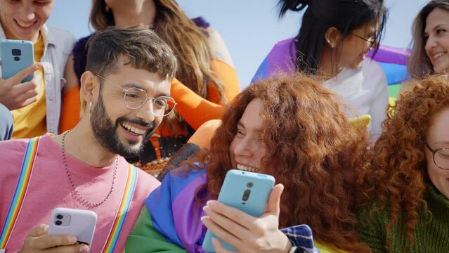 Large multiracial group young friends using cell phones outdoors. LGBT diverse community mobile addicts checking social media at gay pride festival. Generation z and crowded events in city.