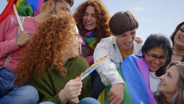 Group multi-ethnic excited people gathered to celebrate gay pride day in street. Diverse joyful friends having fun together sitting at LGBT party with rainbow flags. Generation z and sexual freedom.