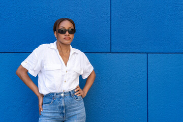 Pretty young woman of black ethnicity with sunglasses looking at the camera and white shirt, on a blue background.