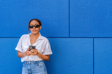 Beautiful young woman of black ethnicity with sunglasses and white shirt looking at the camera with a mobile phone in her hand on social networks, with a blue background.