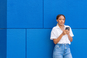 Beautiful young woman of black ethnicity with a white shirt looking at the camera with a mobile phone in her hand on social networks, with a blue background.