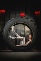 Young beautiful woman in futuristic silver wear is posing in tractor tyre on iron wall background.