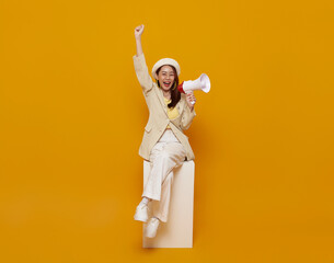 Happy smiling young pretty Asian woman sitting on white box and holding megaphone making announcement isolated yellow studio background.