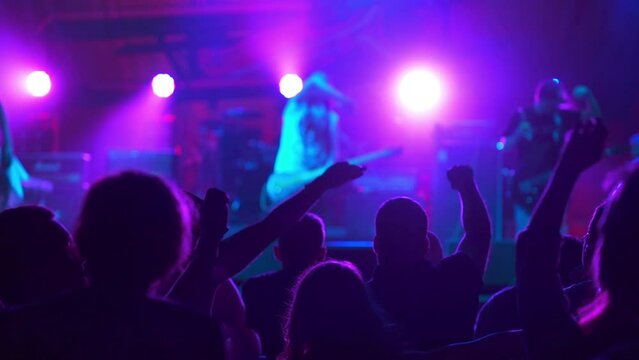Rock Band with Guitarists and Drummer Performing at a Concert in a Night Club. Front Row Crowd is Partying. Silhouettes of Fans Raise and wave Hands in Front of Bright Colorful Strobing Light on Stage