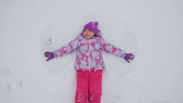 Happy little girl lying on a snow and shows angel, slow motion