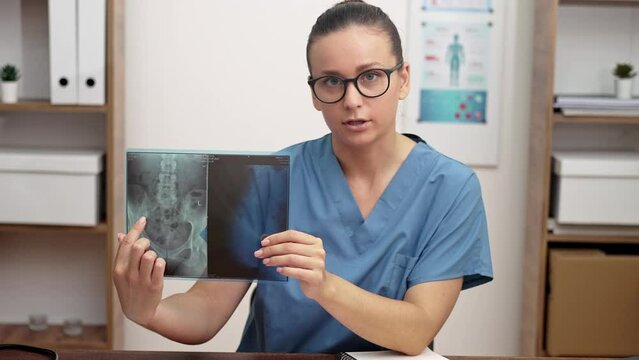 Doctor shows X-ray picture looking at camera sitting in office female in blue lab coat films lesson to students explaining results of patient examination online education
