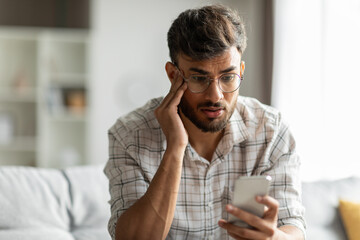 Shocked indian guy looking at cellphone screen, sitting on couch at home, holding phone and touching his head, reading bad news or message, copy space