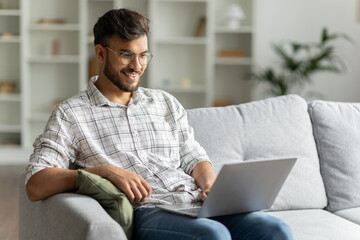 Positive hindu man freelancer working using laptop computer sitting on couch at home interior, guy...