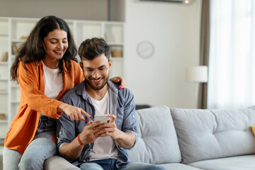 Happy young indian couple using smartphone, wife pointing at cellphone screen, sitting on sofa in...