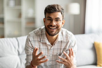 Cheerful indian guy talking looking at camera and gesturing, communicating online sitting on couch...