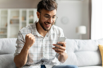 Yes, great news. Overjoyed indian guy holding smartphone and shaking clenched fist, sitting on...