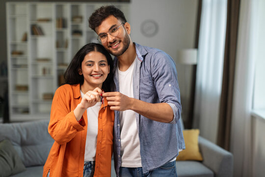 Excited young indian husband and wife showing keys from their new apartment, enjoy buying own home, standing in room interior, copy space. Mortgage real estate concept