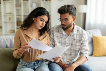 Indian couple checking bills, reading documents, unhappy man and woman holding papers, counting monthly spendings, sitting on sofa at home - obrazy, fototapety, plakaty