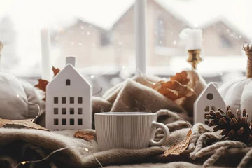  Cozy Autumn. Warm cup of tea, stylish pumpkin pillows, fall leaves, candle, lights and cute buildings decoration on brown scarf on windowsill. Autumn hygge, fall home decor © sonyachny