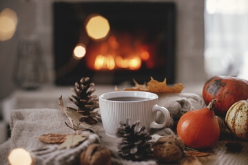 Warm cup of tea, pumpkins, autumn leaves, cozy scarf on rustic wooden table on background of burning fireplace. Hygge fall home, rural banner. Autumn still life. Happy Thanksgiving