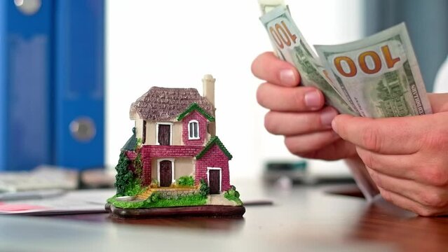 Man counting paper dollar currency with house figurine in front. Concept of renting a house or paying tax for an apartment. First payment mortgage loan
