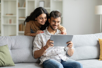 Happy indian man and woman sitting on couch, hugging and using digital tablet, looking at pad screen and smiling, using newest gadget at home interior - obrazy, fototapety, plakaty