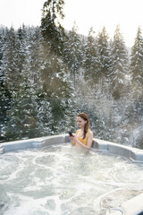 Vertical image of a young woman scrolling a mobile phone while relaxing in a hot jacuzzi bath on a background of snowy trees in winter.