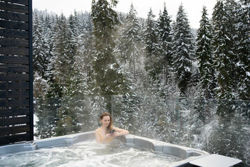 A young woman looks at the phone while relaxing in a hot tub against the background of Christmas trees in winter. The concept of recovery in a thermal spa