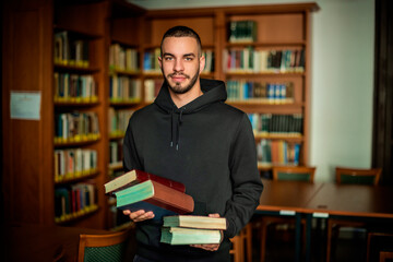 Fototapeta Young man standing in the university library and holding books in his hand obraz