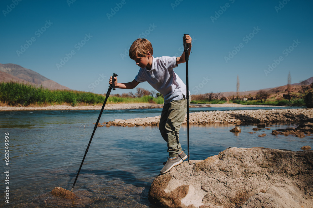 Sticker Boy with trekking poles checks the depth of mountain river