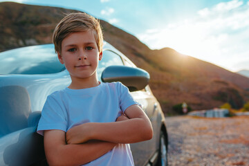 Cut boy stands by the car in the mountains at sunset, travel concept