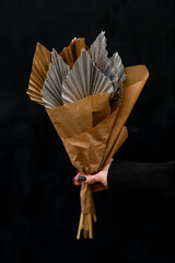 Bouquet of dried leaves in silver and gold colors wrapped in kraft paper in female hand on black background